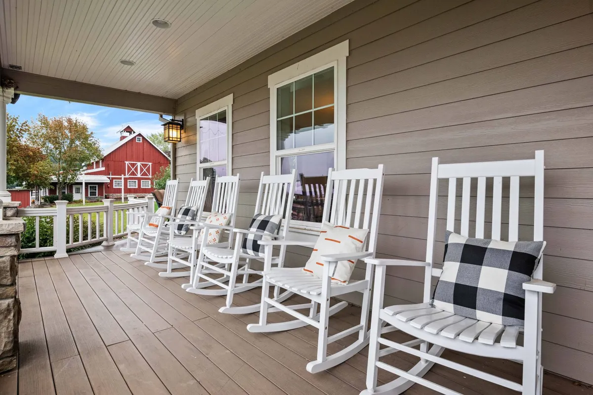 The farmhouse porch with classic rocking chairs overlooks the rolling landscape and distant barn, bathed in warm sunset light. The scene evokes a welcoming, relaxing ambiance, offering guests a chance to unwind while connecting with the historic setting.