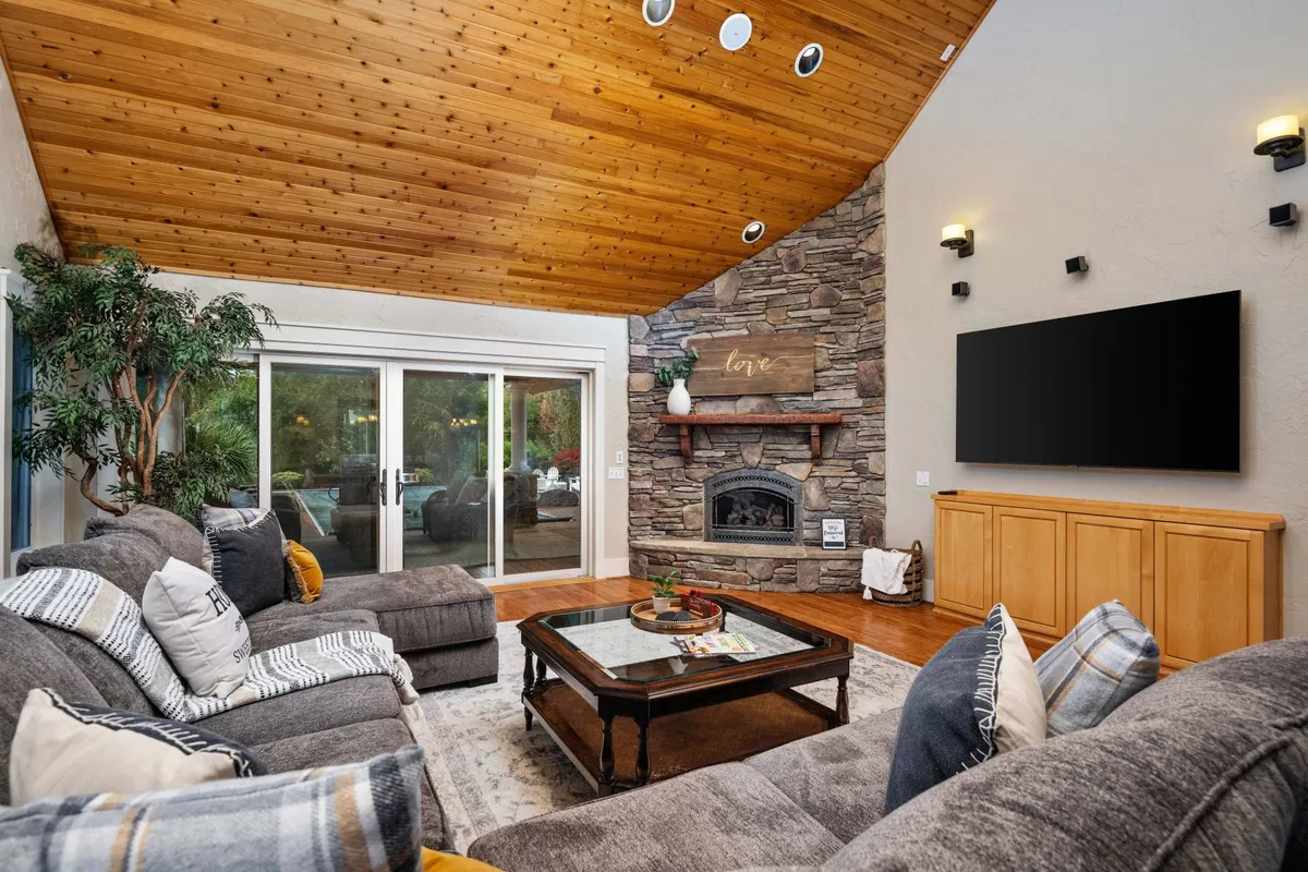 A large, cozy living room featuring a stone fireplace, expansive windows offering views of the outdoors, and comfortable sofas arranged for group conversations or relaxation. A coffee table sits at the center, and the room is awash in natural light, creating an inviting social space.