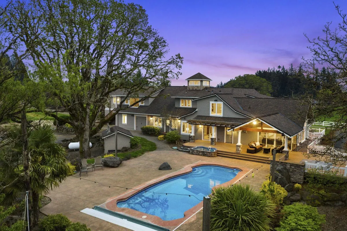 A beautifully landscaped outdoor area featuring a sparkling swimming pool surrounded by comfortable seating and lush gardens. A large tree offers shade, while the inviting pool is perfect for summer relaxation and gatherings. The view extends to open lawns and hints of rural scenery beyond.