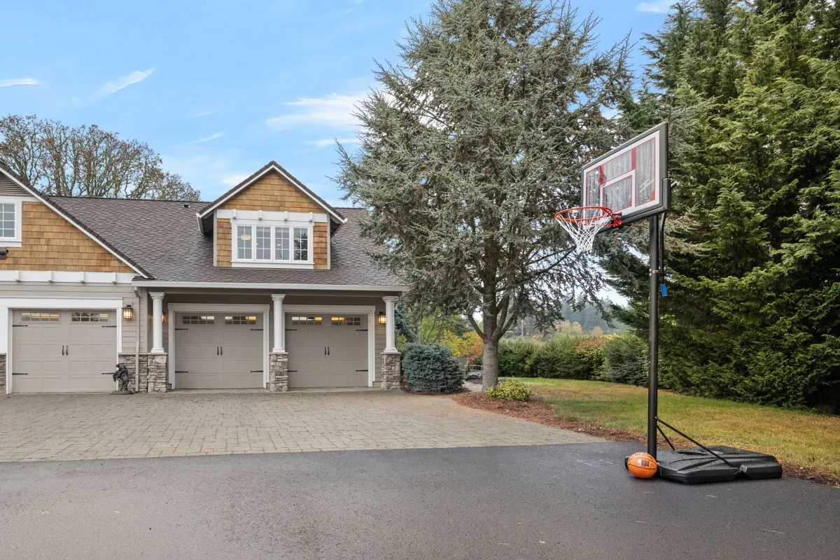 The exterior features a basketball hoop in the driveway, garage doors, and tall trees in the background. The scene evokes playful, active moments and opportunities for outdoor games and bonding.