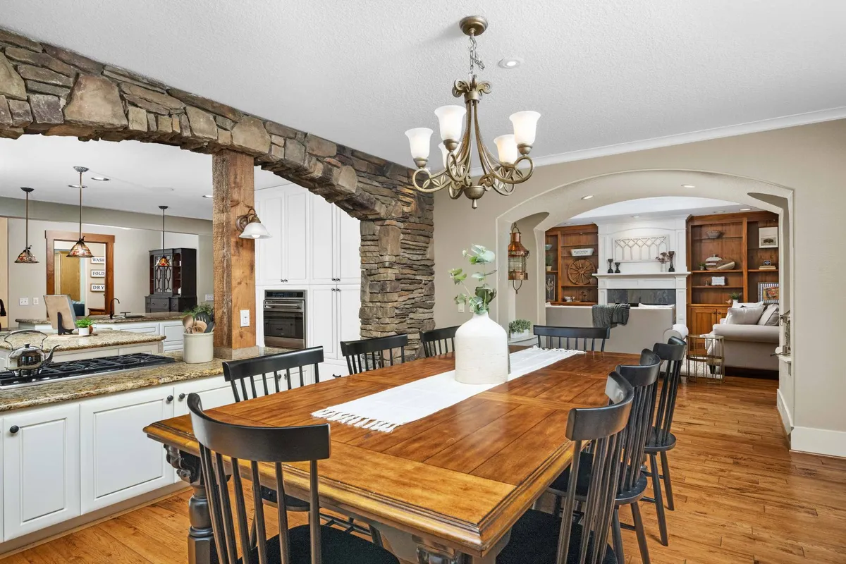 A warm dining space with a wooden table under a chandelier, adjacent to a welcoming kitchen. The stone archway and cozy lighting create an inviting atmosphere, ideal for family meals and celebrations.