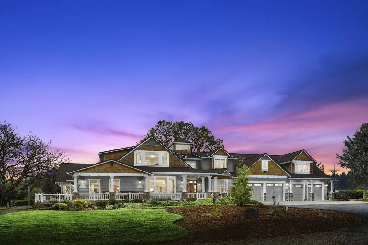 The hero image showcases the grand farmhouse exterior framed by a sprawling, manicured lawn, a spacious front porch perfect for gathering, and inviting architectural details that exude warmth and comfort. The setting is open and sunlit, hinting at the tranquil northwest countryside just beyond the home.