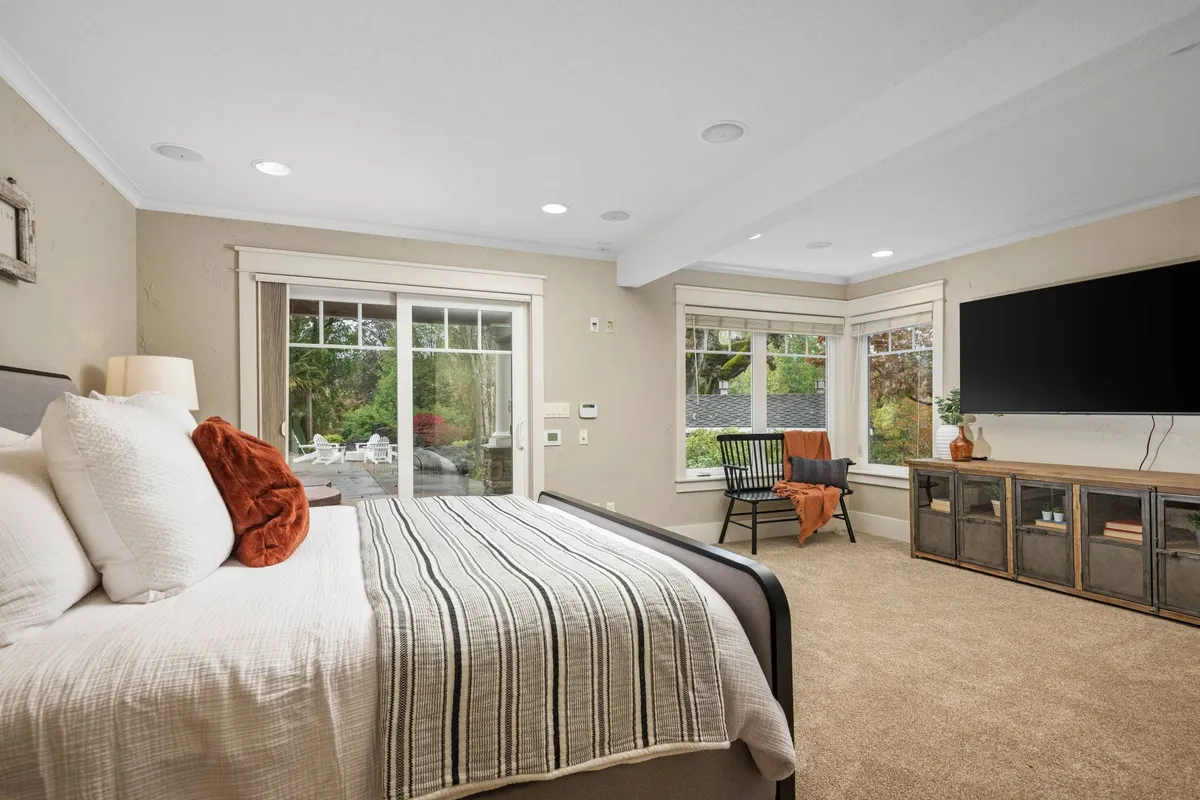 A serene bedroom with a bed adorned with decorative pillows, large windows offering tranquil views, a modern wall-mounted TV, and a stylish side table. Natural light floods the room, creating a calming atmosphere ideal for relaxation and restful nights.