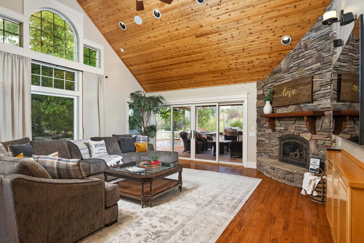 A cozy living area with a large sectional sofa, a striking stone fireplace, wooden ceilings, and expansive windows offering panoramic views of rolling hills.
