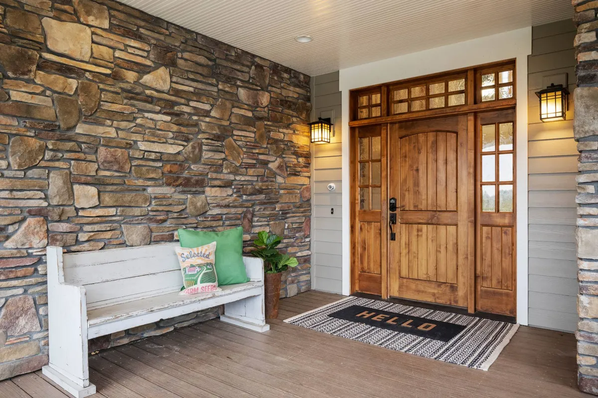The photograph captures the welcoming front entrance of the farmhouse, featuring a sturdy wooden door set in a stone wall, a cozy bench with pillows, and a homey welcome mat. The scene is bathed in natural light, giving a sense of arrival and warmth.